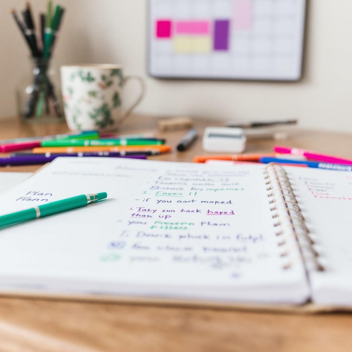 A colorful desk scene with a planner, handwritten notes, and art supplies. A wall calendar shows highlighted tasks, depicting a person organizing their week manually.